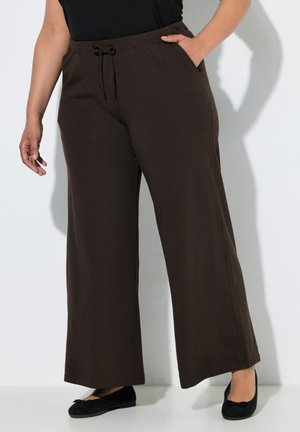Personne portant un pantalon large marron foncé avec une taille élastique à cordon, debout avec une main dans la poche et des chaussures plates noires.