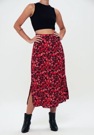Kobieta ubrana w czarny bezrękawnik typu crop top, czerwoną i czarną kwiecistą spódnicę midi z bocznym rozcięciem, czarne botki i złotą bransoletkę na lewej ręce.