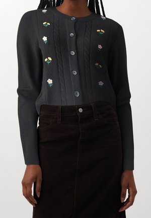Zwarte gebreide cardigan met kabelpatroon en bloem borduursels; beschikt over een ronde halslijn en knoopsluiting, gedragen met een bruine corduroy rok.