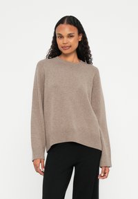 Bež pleten sweater s sproščenim krojem, okroglim izrezom in rebrastimi manšetami ter robom, v kombinaciji s črnimi hlačami. Gladka tekstura.
