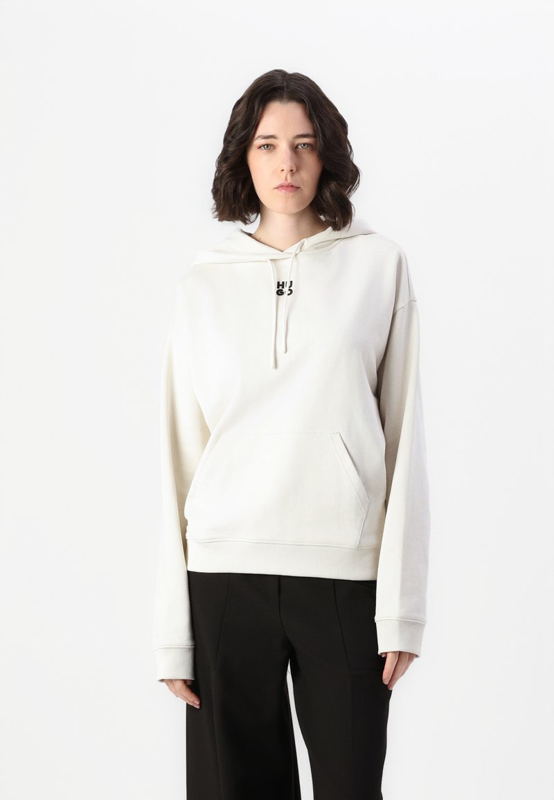Felpa leggera beige con tasca frontale e cappuccio regolabile con cordino. Presenta un piccolo logo nero sul petto. Abbinata a pantaloni neri a gamba larga.