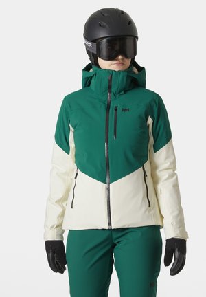 Grüne und cremefarbene Skijacke mit einem Reißverschluss vorne, hohem Kragen und verstellbarem Kapuze. Glänzende schwarze Handschuhe runden das Outfit ab.