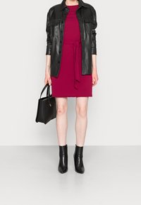 Schwarze Lederhemdjacke über einem burgunderfarbenen Kleid mit einem Bund an der Taille, kombiniert mit schwarzen Knöchelstiefeln und einer schwarzen Tragetasche.