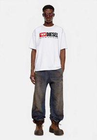 Bela oversized majica z rdečim in črnim logotipom "DIESEL", kombinirana z ohlapnimi, zbledelimi modrimi kavbojkami in rjavimi delovnimi škornji.