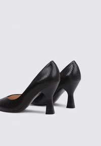 Zapatos de tacón alto de charol negro con un acabado texturizado, punta redondeada y tacones cónicos de 3 pulgadas. De pie sobre un fondo neutro.