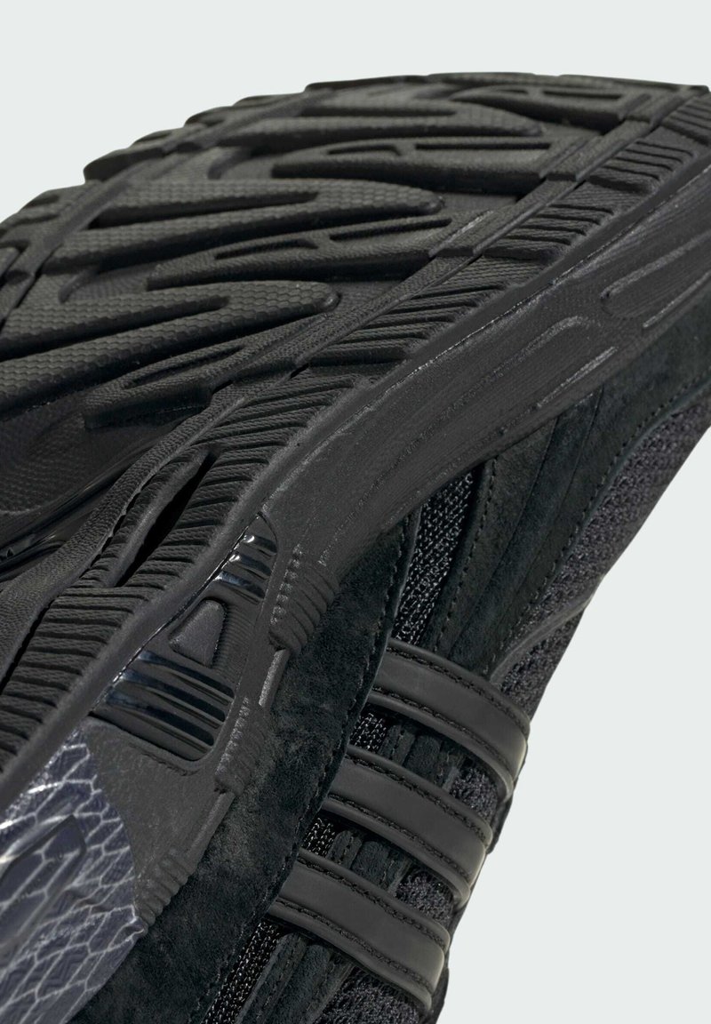 Suola di scarpa atletica nera in gomma testurizzata, con design curvo, piccole scanalature e tre strisce orizzontali lungo il lato.