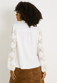 Blouse blanche à manches longues en dentelle avec des motifs délicats, un col rond et une coupe décontractée au dos.