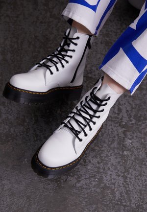 Bottes blanches en cuir à lacets avec semelles noires et coutures jaunes, portées avec un pantalon blanc arborant de larges motifs géométriques bleus.