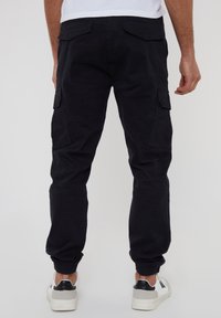 Černé pracovní kalhoty s elastickými manžetami na kotnících, vybavené několika bočními kapsami a hladkou bavlněnou texturou. Pohled zezadu ukazuje dvě zadní kapsy.