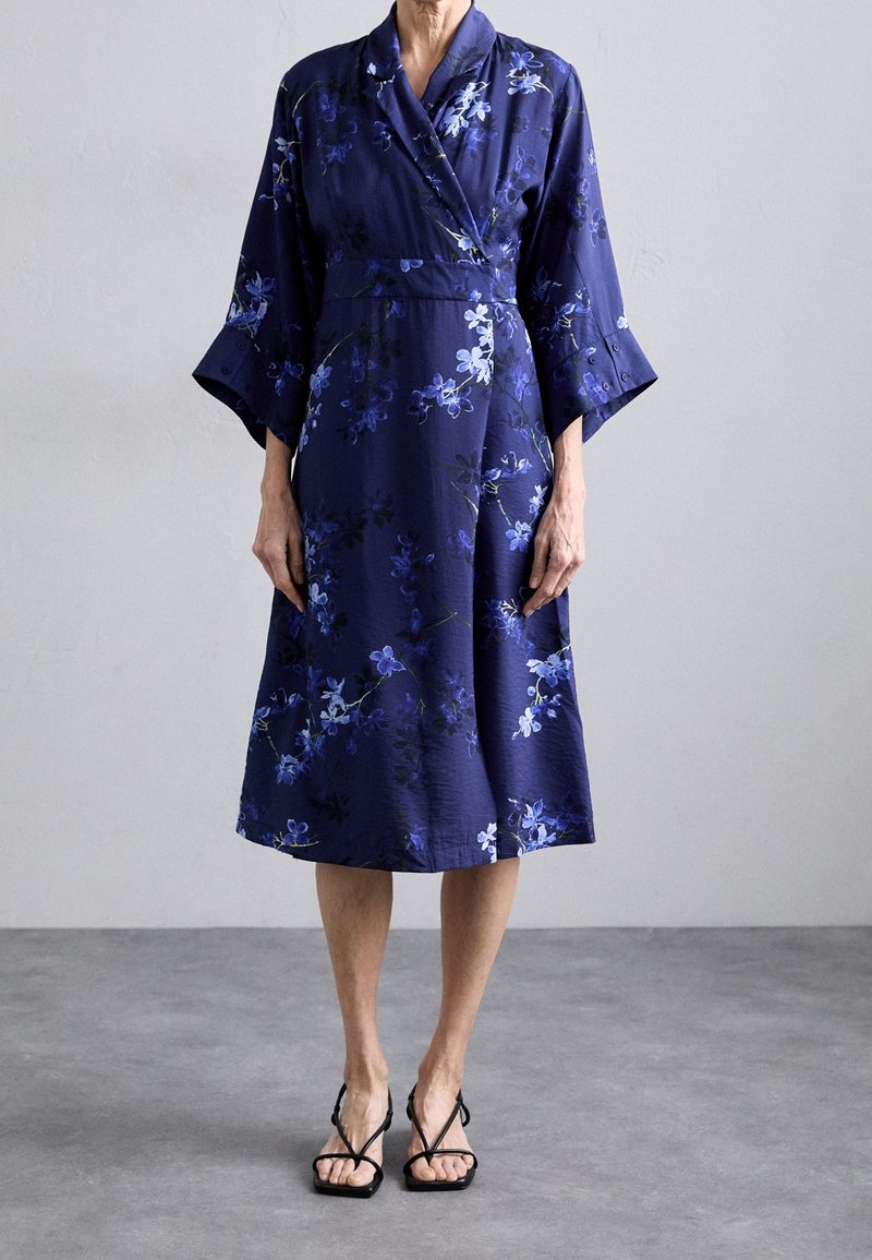 Femme portant une robe portefeuille fleurie bleu marine jusqu'au genou avec des manches larges et des sandales noires à brides, debout sur un sol gris.