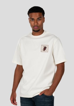 Camiseta de algodón blanca con un parche bordado cuadrado que presenta un diseño de personaje, cuello redondo y mangas cortas. Combinada con jeans oscuros.