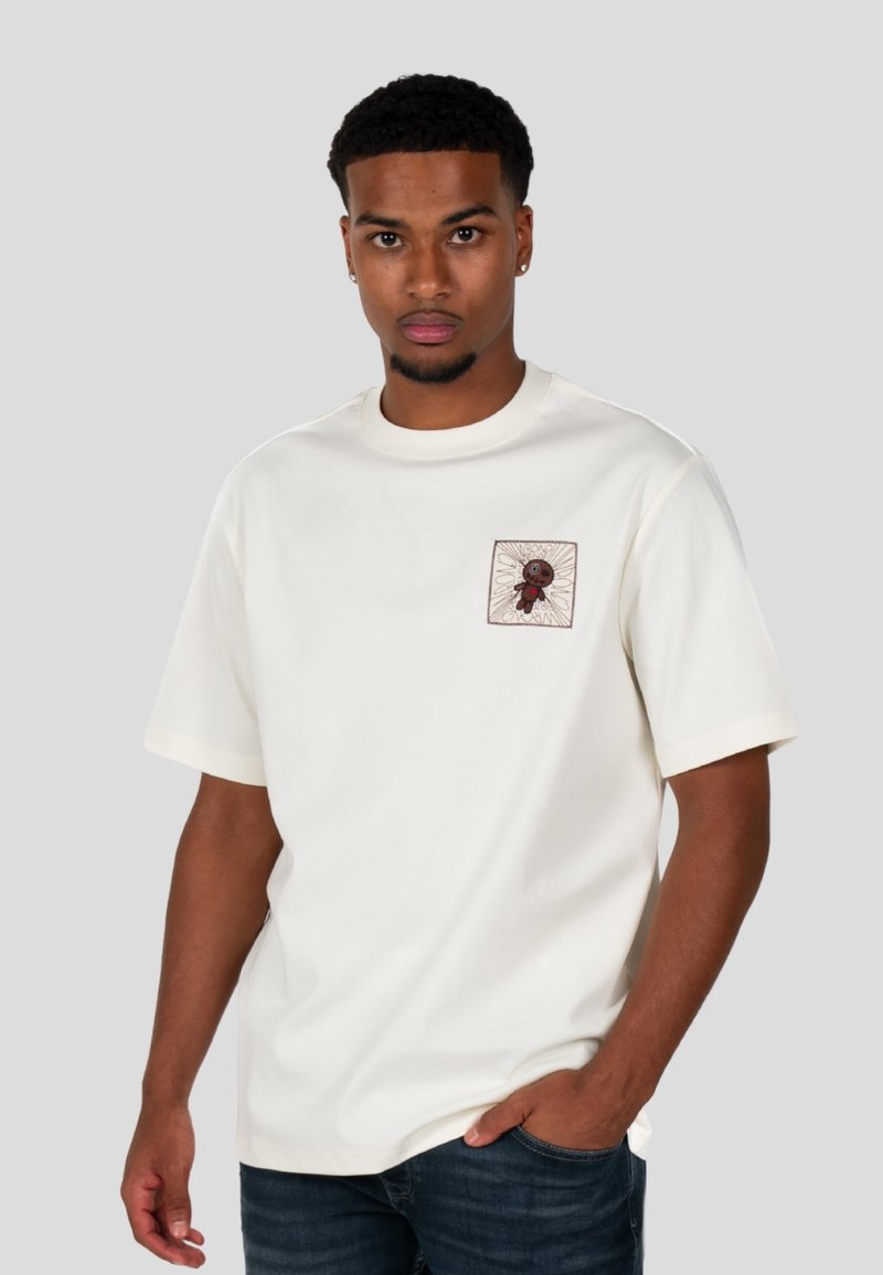 Bílá bavlněná trička s vyšitým čtvercovým nášivkem s designem postavy, kulatým výstřihem a krátkými rukávy. Nosené s tmavými džíny.