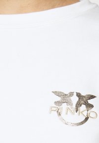 Valkoinen puuvillapaita, jossa on kultainen metallinen logo, jossa on kaksi lintusilhuettia ja teksti "PINKO" alla. Sileä pinta, pyöreä kaula-aukko.