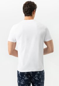 Man met kort, krullend haar in een eenvoudig wit shirt met korte mouwen en marineblauwe shorts met witte cartoonfiguren, gezien van achteren.