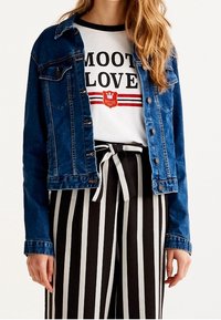 Femme portant une veste en jean bleue, un t-shirt blanc graphique avec le texte "MOOT LOVE", et un pantalon taille haute à rayures verticales noires et blanches avec une ceinture nouée.