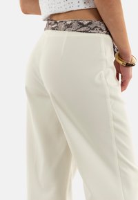 Pantaloni bianchi a gamba larga con una texture liscia, caratterizzati da una cintura con stampa a serpente e tasche laterali. Orologio dorato al polso.