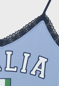 Camisole bleu clair avec bordure en dentelle noire, bretelle marine et lettres blanches en bloc épelant partiellement « ALIA » au-dessus d’un détail de drapeau vert et blanc.