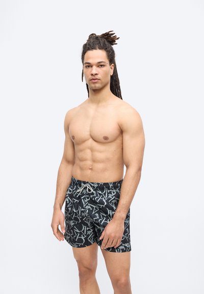 Μυώδης άνδρας με μακριά dreadlocks, που φοράει μαύρο μαγιό με σχέδια, στέκεται μπροστά σε απλό φωτεινό φόντο.