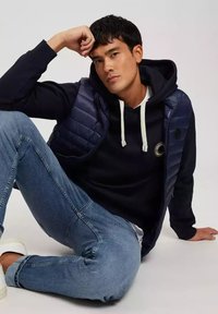 Granatowa ocieplana kamizelka na czarnej bluzie z kapturem, połączona z jasnoniebieskimi dżinsami i białymi trampkami, siedząca na neutralnym tle.