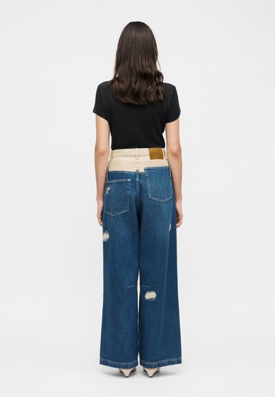 Femme aux longs cheveux foncés portant un haut noir à manches courtes et un jean taille haute à jambes larges avec une ceinture beige et des zones usées, debout de dos.