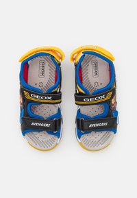 Zilās un melnās sandales ar dzelteniem akcentiem, Velcro siksnām, perforētu zoli un Avengers grafiku, kas izstrādātas ventilācijai un komfortam.