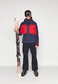 Skijaška oprema koja uključuje tamnoplavu i crvenu jaknu otpornu na vremenske uvjete s kapuljačom, crne hlače i skije s vezovima, prikazane u uspravnom položaju.