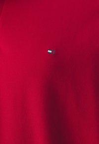 Červené polo tričko vyrobené z texturované látky, které má na levé části hrudi malé vyšívané logo v modré, bílé a černé barvě.