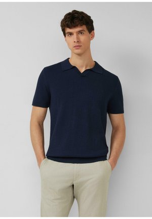 Mężczyzna stoi z rękami w kieszeniach, ubrany w granatową, krótkozręczną dzianą koszulkę polo i jasnobeżowe spodnie na szarym tle.