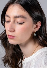 Orecchino a cerchio dorato con un cristallo a forma di fiore di colore azzurro chiaro. La modella ha i capelli lunghi e scuri e indossa un top bianco con una texture e un scollo semplice.