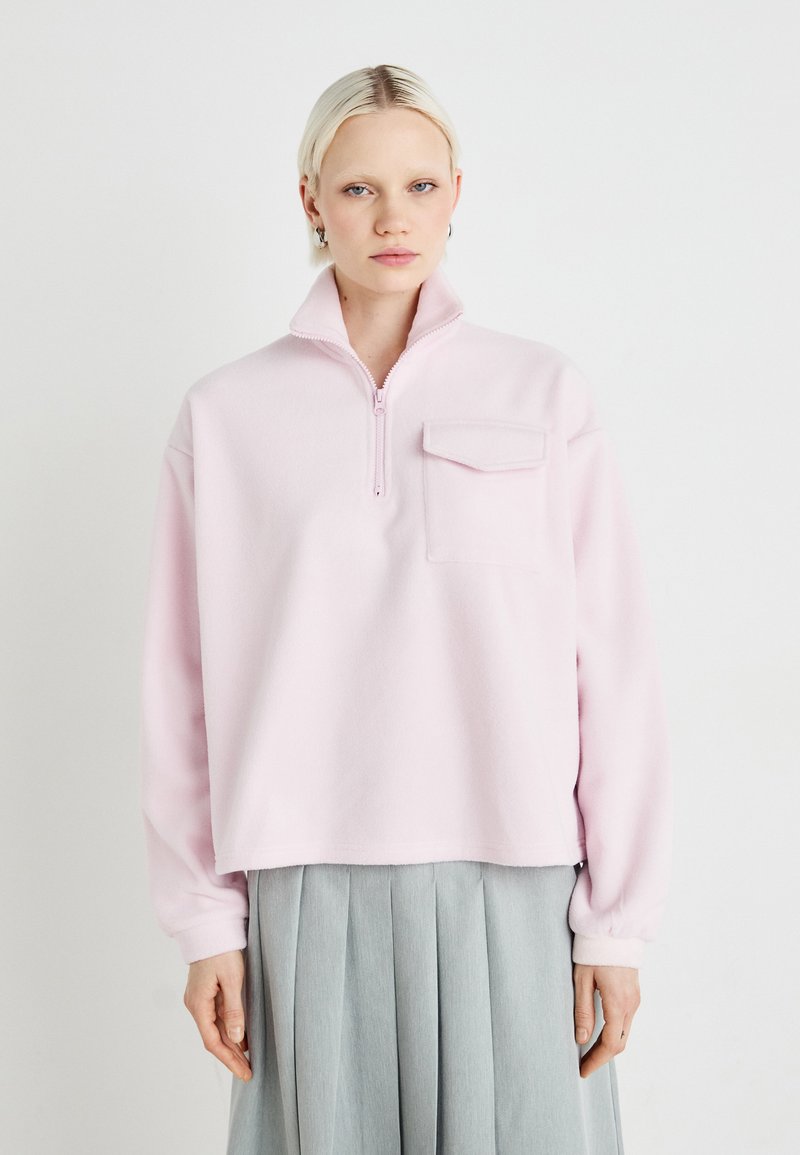 Lichtroze fleece pullover met een zijdezachte structuur, een ritssluiting bij de hals, lange mouwen en een borstzak. De stof heeft een zachte textuur en een comfortabele pasvorm.