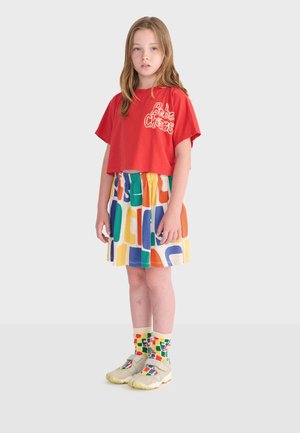 Jeune fille debout portant un t-shirt court rouge, une jupe à motif abstrait multicolore, des chaussettes assorties et des chaussures beige clair sur un fond uni.
