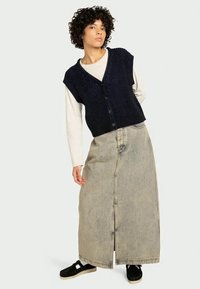 Colete canelado azul-marinho com botões, usado sobre uma camisa de mangas longas cremosa, combinado com uma saia longa de ganga bege e sapatos pretos deslizantes.