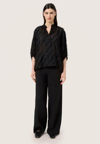 Blusa nera con texture, dal taglio ampio e oversize, con mezza abbottonatura, design a strati e maniche tagliate fino al gomito, abbinata a pantaloni larghi.
