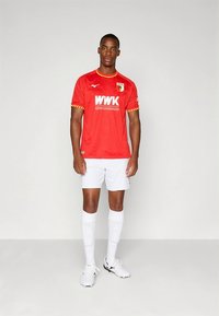 Červený fotbalový dres s krátkými rukávy, žlutými akcenty a logem, doplněný bílými šortkami a bílé ponožky po kolena.