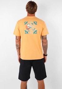 T-shirt en coton jaune affichant un graphique floral et de feuilles en bleu et orange au dos. Manches courtes et coupe décontractée. Shorts noirs portés.