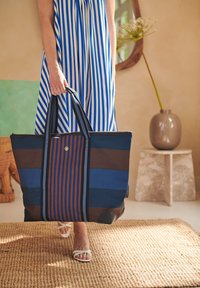Gestreepte tote bag in blauw en bruin met dubbele handvatten, textuurstof, gouden accentknop en zwarte onderste hoeken. Geweven tapijt eronder.