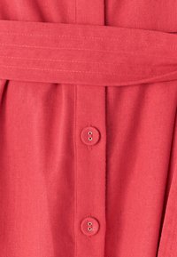 Korálové šaty z látky s opaskom v páse, ktoré majú zapínanie na gombíky a jemnú textúru. Sú viditeľné dva okrúhle gombíky s očkami.