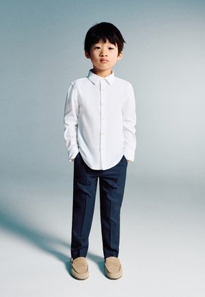 Junger Junge steht mit den Händen in den Taschen, trägt ein weißes Hemd mit Knöpfen, marineblaue Hose und beige Schlupfschuhe vor einem einfarbigen Hintergrund.