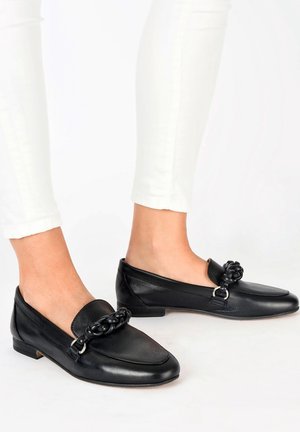 Mocasines de cuero negro con un acento de cadena texturizada en el empeine, tacón plano y punta redondeada. Llevados con pantalones blancos ajustados.