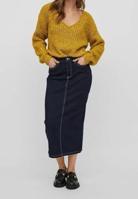 Senfgelb gestrickter Pullover mit V-Ausschnitt und lockerer Passform, kombiniert mit einem dunkelblauen Denim-Maxikleid featuring weißen Nähten und leichtem Schlag am Saum.