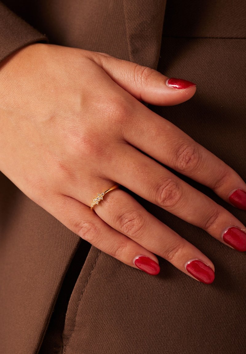 Gouden ring met kleine, heldere stenen in een golvend patroon. Hand rustend op een bruine stoffen ondergrond. Rode nagellak op de vingers.