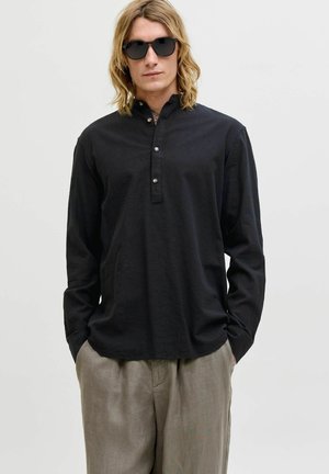 Mees, kellel on õlgadeni ulatuvad blondid juuksed, mustad päikeseprillid, must pikkade varrukatega nööbitud särk ja beežid laiad püksid, käed taskutes.