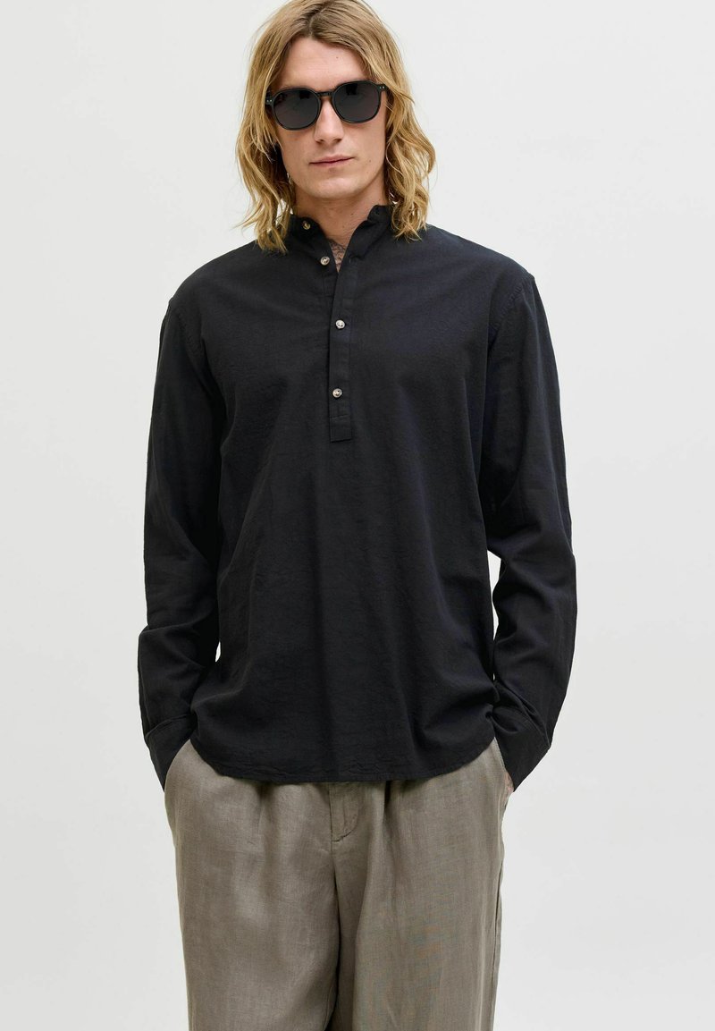 Homme aux cheveux blonds mi-longs portant des lunettes de soleil noires, une chemise noire à manches longues boutonnée, et un pantalon beige ample, les mains dans les poches.