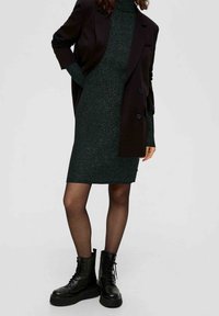 Personne portant une robe en maille verte foncée, un blazer noir, un collant noir transparent et des bottes compensées noires à lacets, debout avec une main sur la hanche.