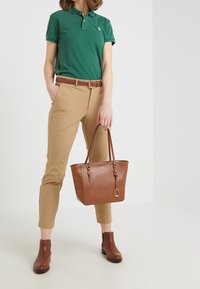 Grön polo skjorta, beige skräddarsydda byxor, bruna läderankelboots och en brun toteväska med gulddetaljer. Strukturerad yta på väskan.