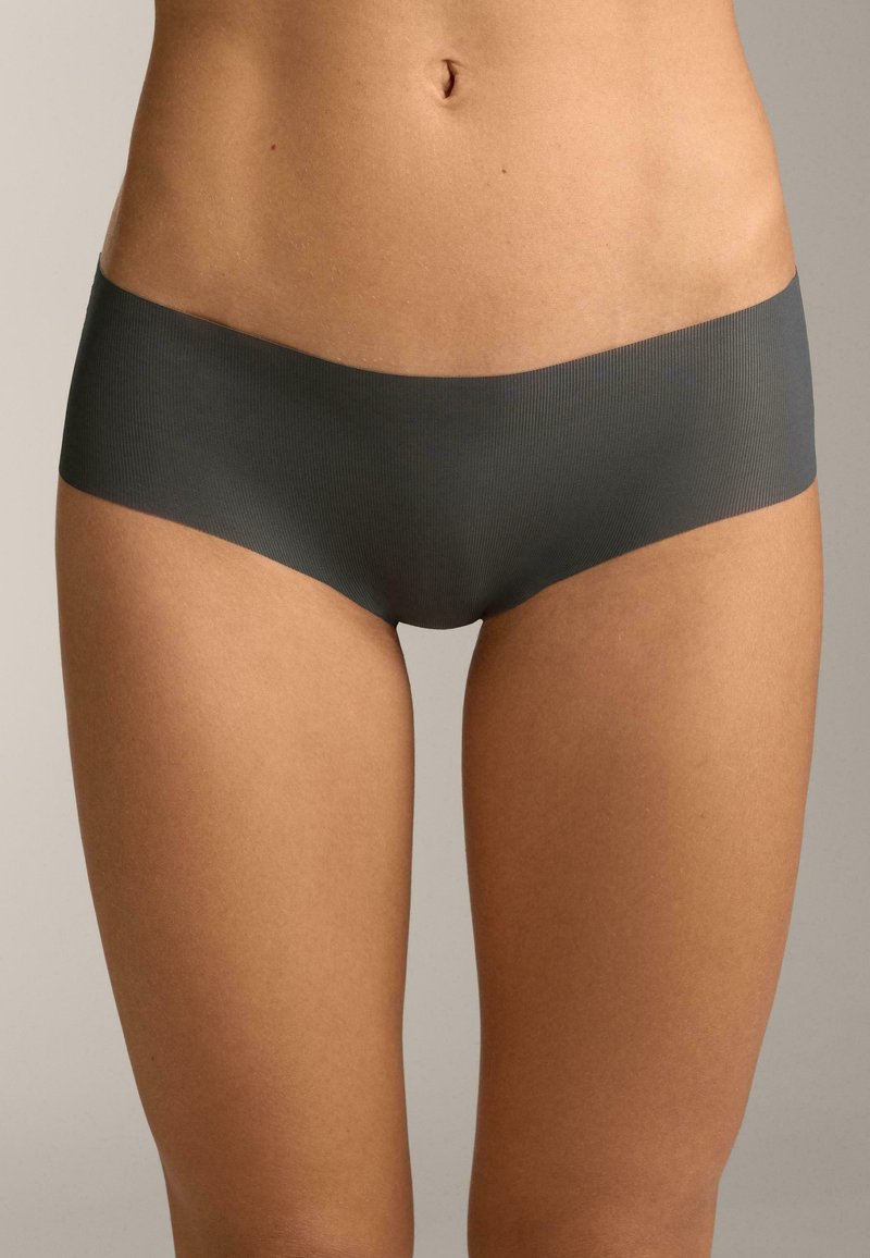 Femme portant une culotte hipster gris foncé sans coutures, debout devant un fond clair uni.