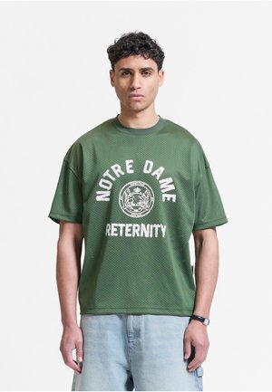 Młody mężczyzna z krótkimi kręconymi włosami, w zielonej koszulce Notre Dame i jasnoniebieskich szortach, patrzy przed siebie z neutralnym wyrazem twarzy.