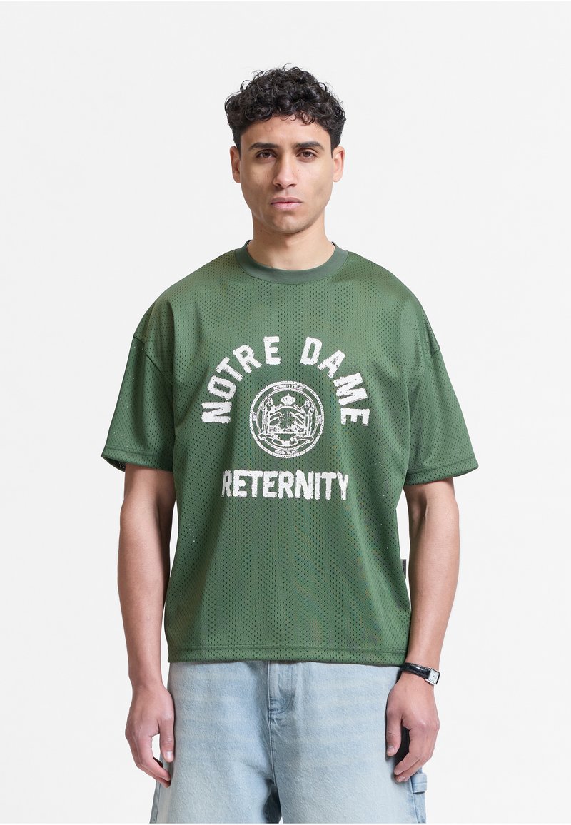 Jeune homme aux cheveux courts et bouclés portant un t-shirt vert Notre Dame et un short bleu clair, regardant vers l'avant avec une expression neutre.