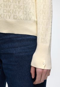 Manche de pull beige DKNY avec bord-côte et petit détail bouton à côté d'un jean en denim bleu foncé sur le bras d'une personne.