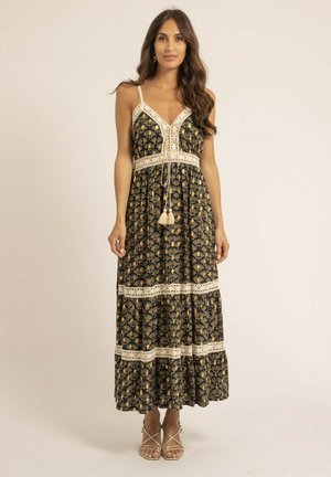 Femme debout portant une robe maxi noire à motifs avec une bordure en dentelle crème et des liens à pompons, chaussant des sandales beiges à lanières sur un fond uni.